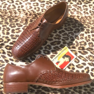 May include: A pair of brown leather dress shoes with woven detailing on the toe and side panels. The shoes have a classic lace-up design and a low heel. A vintage card with the text "LA DOUCHKA VINTAGE" is also present.