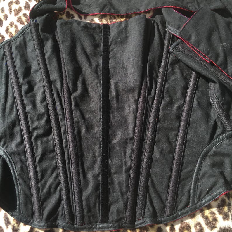 May include: Black corset with vertical boning and red trim. The corset is made of a quilted fabric and features a structured design. The edges are finished with red piping. The overall style is gothic and vintage.