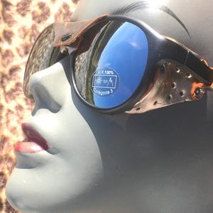 May include: Black sunglasses with blue reflective lenses and brown leather accents. The sunglasses have a unique design with a vintage aesthetic. The lenses have a sticker that reads "U.V. 100%" and "Catégorie 3".