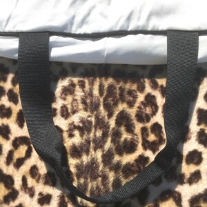 May include: A tote bag with a leopard print pattern. The bag has a black handle and a light gray top. The leopard print is brown and tan. The bag is made of a soft, plush material.