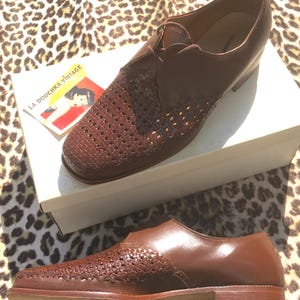 May include: A pair of brown leather shoes with woven detailing on the toe and side panels. The shoes are displayed on a white box with a vintage label that reads "LA DOUCHKA VINTAGE". The shoes have a classic, formal design.