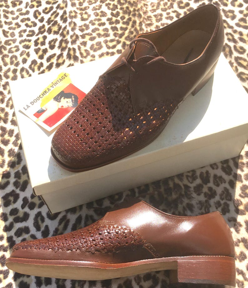 May include: A pair of brown leather shoes with woven detailing on the toe and side panels. The shoes are displayed on a white box with a vintage label that reads "LA DOUCHKA VINTAGE". The shoes have a classic, formal design.