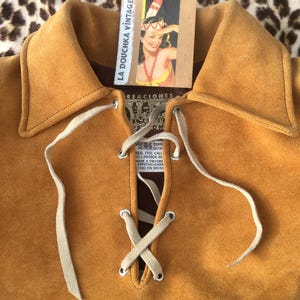Puede incluir: Camisa de ante color mostaza con escote con cordones y cuello. La camisa tiene una etiqueta que dice "LA DOUCHKA VINTAGE" y presenta una ilustraci&oacute;n de chica pin-up vintage. La camisa se muestra sobre un fondo con estampado de leopardo.