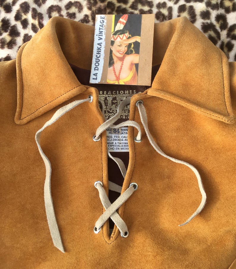 Puede incluir: Camisa de ante color mostaza con escote con cordones y cuello. La camisa tiene una etiqueta que dice "LA DOUCHKA VINTAGE" y presenta una ilustraci&oacute;n de chica pin-up vintage. La camisa se muestra sobre un fondo con estampado de leopardo.