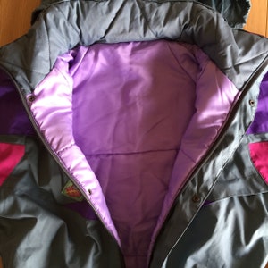 May include: A gray, purple, and pink ski jacket with a purple lining and a small logo patch on the left side. The jacket is unzipped, showing the lining.