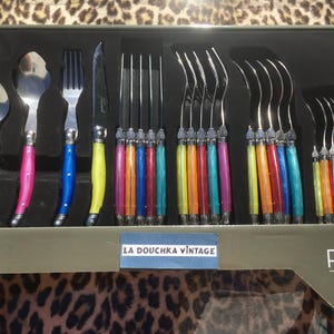 May include: A 24-piece Pradel Heritage cutlery set in a box labelled La Douchka Vintage. The set includes stainless steel spoons, forks, and knives with colourful, translucent, marbled-effect handles in shades of blue, pink, yellow, orange, and purple.