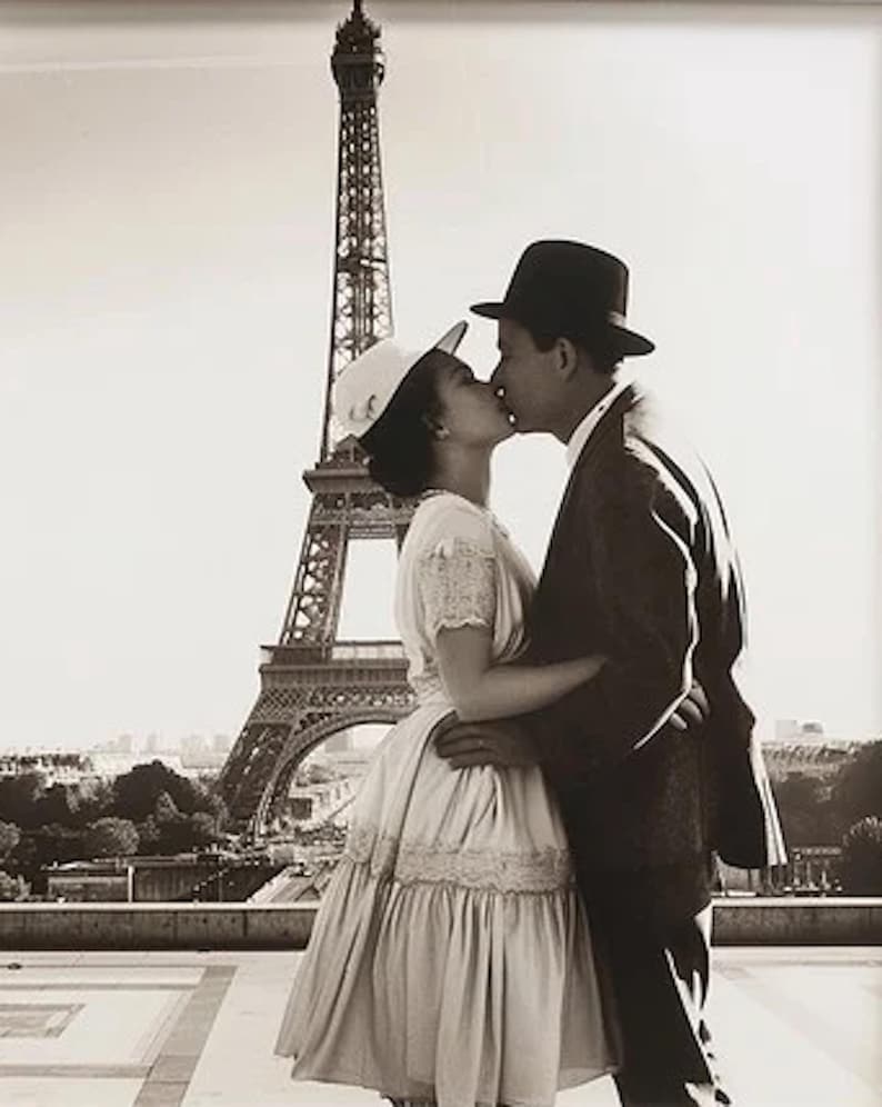 May include: A black and white photograph of a couple kissing in front of the Eiffel Tower. The woman wears a white dress and hat, while the man is in a suit and hat. The iconic Parisian landmark is in the background.