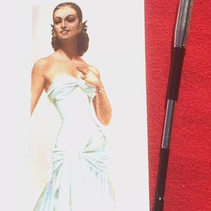 May include: An advertisement for "LA DOUCHKA VINTAGE" featuring a vintage illustration of a woman in a white dress. A black and silver pen is positioned on a red background. The pen has a silver tip and a black body.