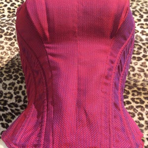 May include: A magenta-colored corset with a textured fabric. The corset has a structured design with vertical seams and a flared bottom. The color transitions to a darker shade of purple along the edges.