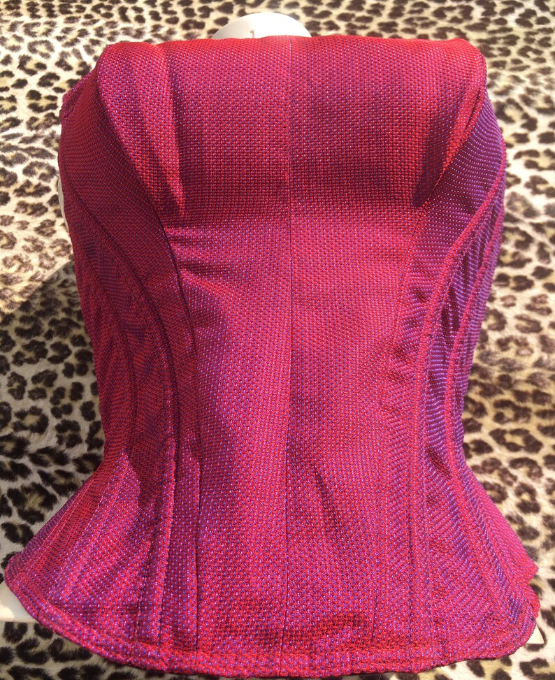 May include: A magenta-colored corset with a textured fabric. The corset has a structured design with vertical seams and a flared bottom. The color transitions to a darker shade of purple along the edges.