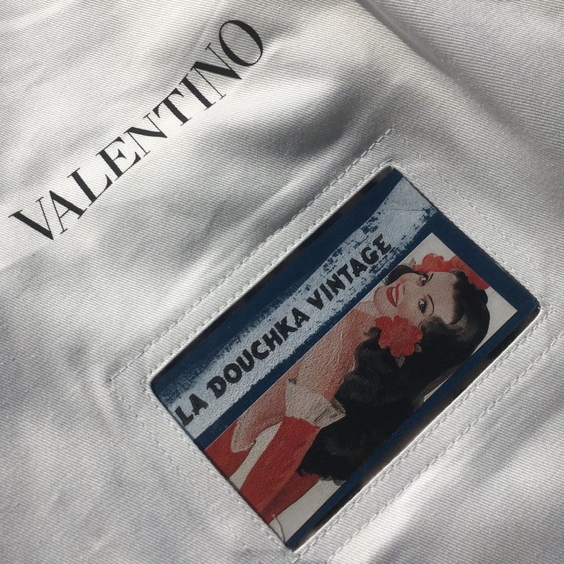 May include: White fabric with the word "VALENTINO" printed in black. A rectangular cutout reveals a vintage-style illustration of a woman with red flowers and the text "LA DOUCHKA VINTAGE".