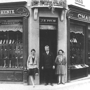 May include: Black and white photograph of a shoe store with the words "AU PHENIX" and "CHAUSSURES" above the windows. Three people stand in front of the store. The windows display various shoe styles.