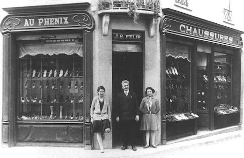 May include: Black and white photograph of a shoe store with the words "AU PHENIX" and "CHAUSSURES" above the windows. Three people stand in front of the store. The windows display various shoe styles.