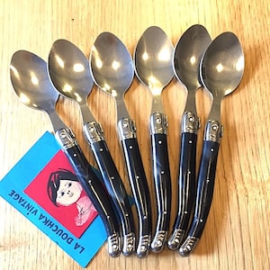 Può includere: Set di sei cucchiai in acciaio inossidabile con manici neri. Ogni cucchiaio presenta un manico curvo, una ghiera argentata e tre punti bianchi. Un'etichetta blu con il testo "LA DOUCHKA VINTAGE" è visibile.