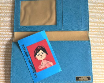 May include: A teal blue leather wallet with a gold label that reads "Laurige Made in France". The wallet has a clear plastic window for a photo ID and a blue and red card with the text "La Doucka Vintage" and a drawing of a woman.
