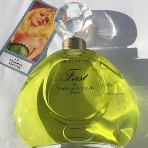 May include: A vintage perfume bottle of "First" by Van Cleef & Arpels, Paris, with a clear, rounded stopper and a light green liquid. The bottle is next to a vintage card with a blonde woman illustration and the text "LA DOUCHKA VINTAGE".