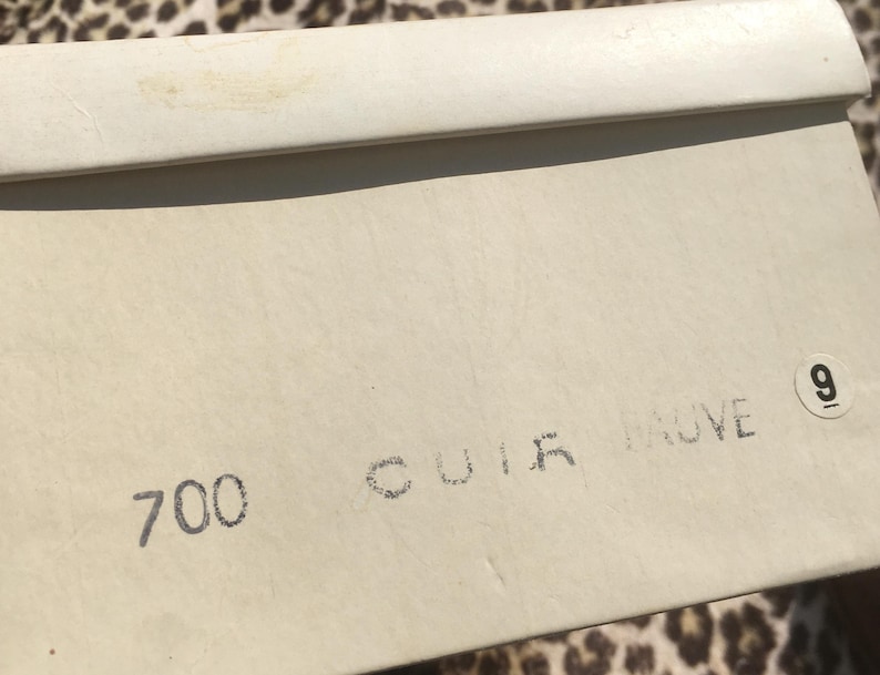 May include: A cream-colored shoe box with the number 9 and the text "700 CUIR TAUVE" printed on the side. The box is made of a textured material.