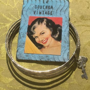 Puede incluir: Un conjunto de pulseras plateadas con un dije de llave, junto a una tarjeta vintage con el texto "LA DOUCHKA VINTAGE" y una ilustración retro de una mujer con lápiz labial rojo.