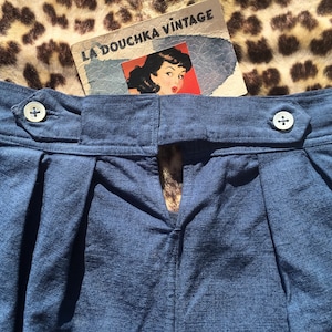 May include: Close-up of blue denim shorts with pleats and two white buttons. The shorts have a high waist and a vintage aesthetic. A vintage tag with the text "LA DOUCHKA VINTAGE" is visible.