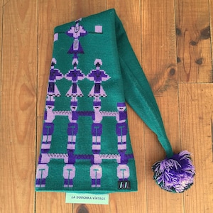 May include: Green knitted hat with a purple and white geometric pattern of figures. The hat has a long, pointed tip and a fluffy purple pom-pom.