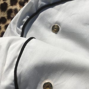 May include: Close-up of a white garment with black trim and gold-colored buttons. The fabric appears to be a textured cotton or linen. The buttons are circular with a detailed design.