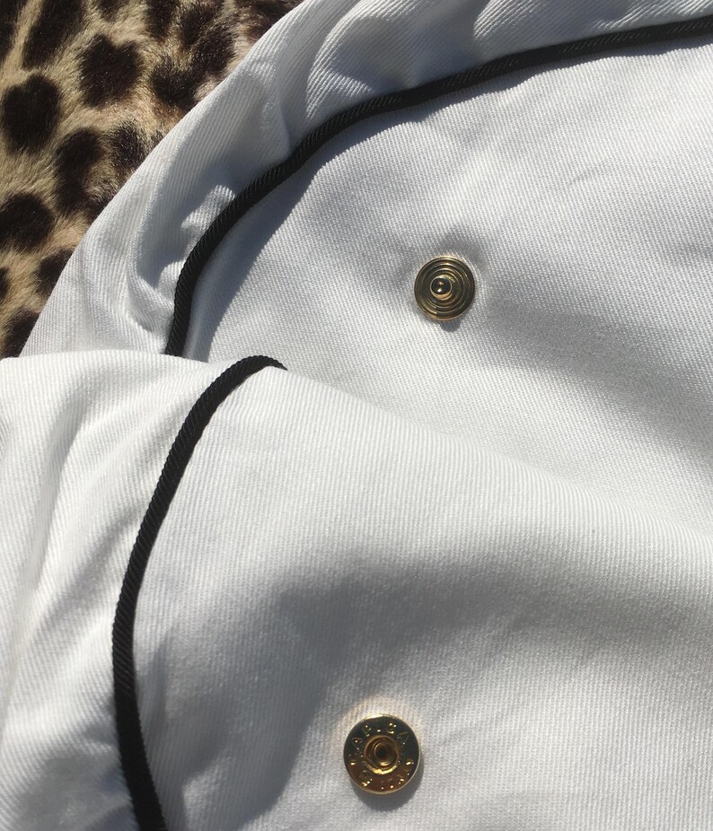 May include: Close-up of a white garment with black trim and gold-colored buttons. The fabric appears to be a textured cotton or linen. The buttons are circular with a detailed design.