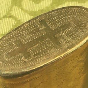 Puede incluir: Un recipiente de metal dorado con una superficie texturizada y un diseño en relieve en la parte superior. El diseño es una cruz con un círculo en el centro.