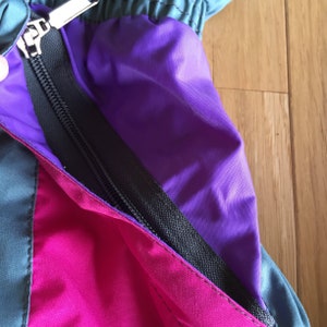 May include: Close-up of a zipper on a jacket with purple, pink, and teal fabric panels.
