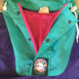 Op de afbeelding: Een teal, paarse en rode K-Way jas met een capuchon. De jas heeft een roze rits en witte trekkoorden. Een patch met het K-Way logo en vlaggen is zichtbaar op het onderste deel van de jas.