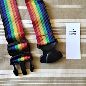 Könnte beinhalten: Ein regenbogenfarbener Gepäckgurt mit schwarzen Plastik-Schnallen. Der Gurt liegt auf einem gestreiften Stoffhintergrund. Eine weiße Karte mit dem Text "LA DOUCHKA VINTAGE" befindet sich im Hintergrund.
