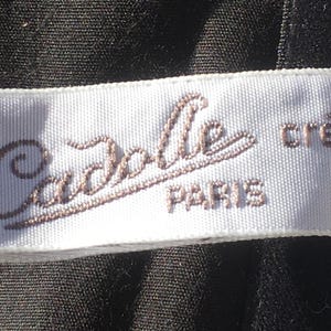 May include: A white label with the text "Cadolle PARIS cr&eacute;ation" in brown script. The label is rectangular and appears to be made of fabric, possibly satin or a similar material. The background is a dark, textured fabric.