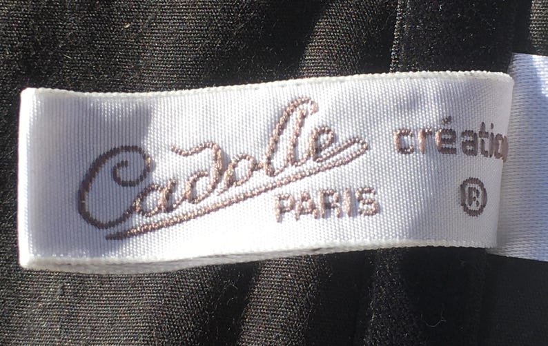 May include: A white label with the text "Cadolle PARIS cr&eacute;ation" in brown script. The label is rectangular and appears to be made of fabric, possibly satin or a similar material. The background is a dark, textured fabric.