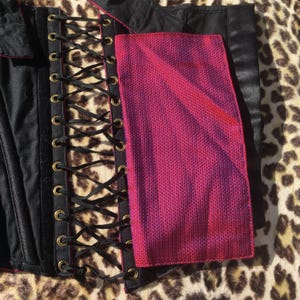 May include: A black corset with a pink and purple panel. The corset features a lace-up front with black ribbon and gold-colored eyelets. The pink and purple panel has a textured appearance.