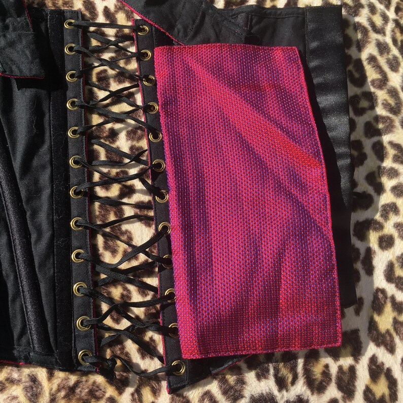 May include: A black corset with a pink and purple panel. The corset features a lace-up front with black ribbon and gold-colored eyelets. The pink and purple panel has a textured appearance.