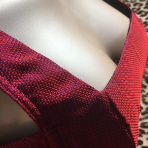 May include: Close-up of a red dress with a textured fabric. The dress has a deep V-neckline and a crisscross design across the chest. The fabric has a subtle sheen, with hints of blue throughout the red material.