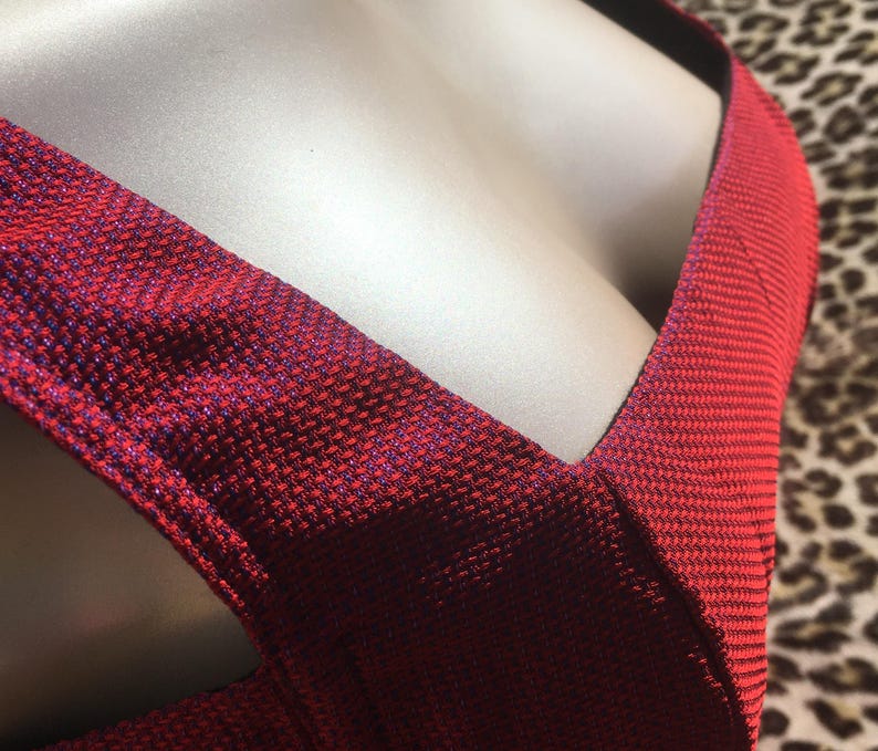 May include: Close-up of a red dress with a textured fabric. The dress has a deep V-neckline and a crisscross design across the chest. The fabric has a subtle sheen, with hints of blue throughout the red material.