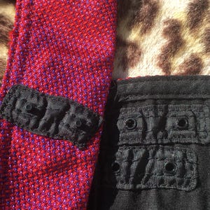 May include: Close-up of a red and purple textured fabric with a black accent piece featuring three small, round, black details. Adjacent is a black fabric with two sets of two round black details.