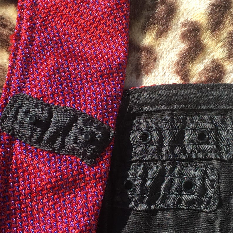 May include: Close-up of a red and purple textured fabric with a black accent piece featuring three small, round, black details. Adjacent is a black fabric with two sets of two round black details.
