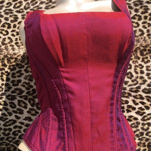 May include: A red and purple corset with a square neckline and structured boning. The corset is displayed on a white mannequin bust with the brand name "AUBADE" visible. The fabric has a textured weave.