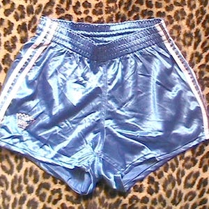 Könnte beinhalten: Glänzende, hellblaue Sporthosen mit weißen Streifen an den Seiten. Die Shorts haben eine hohe Taille und ein kleines Logo am linken Bein. Der Stoff hat einen satinartigen Glanz.