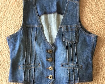 Op de afbeelding: Een blauwe denim vest met een V-hals, vijf knopen en twee zakken. De vest heeft plooien aan de voor- en achterkant.