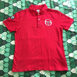 Könnte beinhalten: Rotes Poloshirt mit einem weißen Logo auf der Brust. Das Logo ist ein Kreis mit einem Buchstaben T darin. Auf dem linken Ärmel befindet sich ein kleiner Aufnäher.