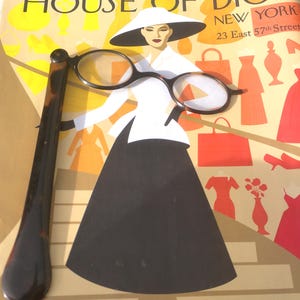 Puede incluir: Un anuncio de House of Dior New York, con una ilustración de moda de una mujer con sombrero blanco y falda negra. Un par de gafas y un mango largo de color oscuro están en primer plano.
