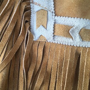 Puede incluir: Primer plano de un art&iacute;culo de ante marr&oacute;n con largas flecos verticales. El borde superior presenta un borde decorativo con detalles blancos y formas geom&eacute;tricas. La textura del ante es visible.