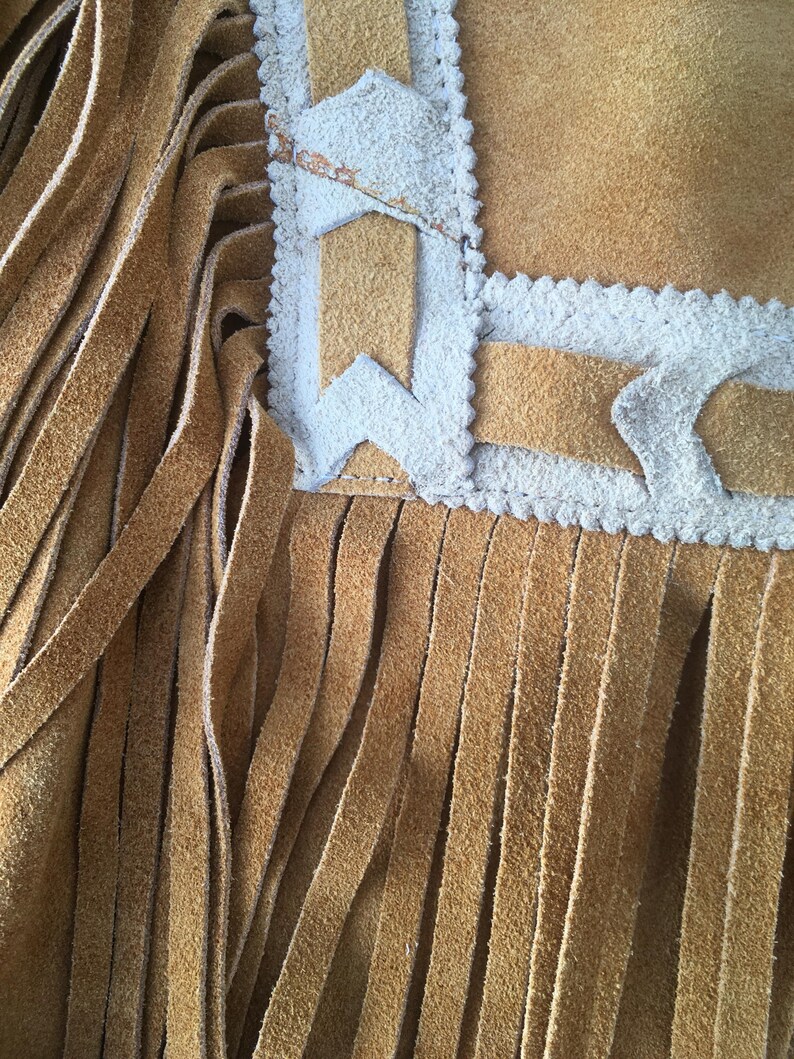 Puede incluir: Primer plano de un art&iacute;culo de ante marr&oacute;n con largas flecos verticales. El borde superior presenta un borde decorativo con detalles blancos y formas geom&eacute;tricas. La textura del ante es visible.