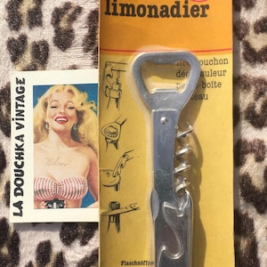 May include: Vintage bottle opener and corkscrew combo on a yellow card with the word "limonadier". The card also features a pin-up style image and the text "LA DOUCHKA VINTAGE". The opener is made in France.