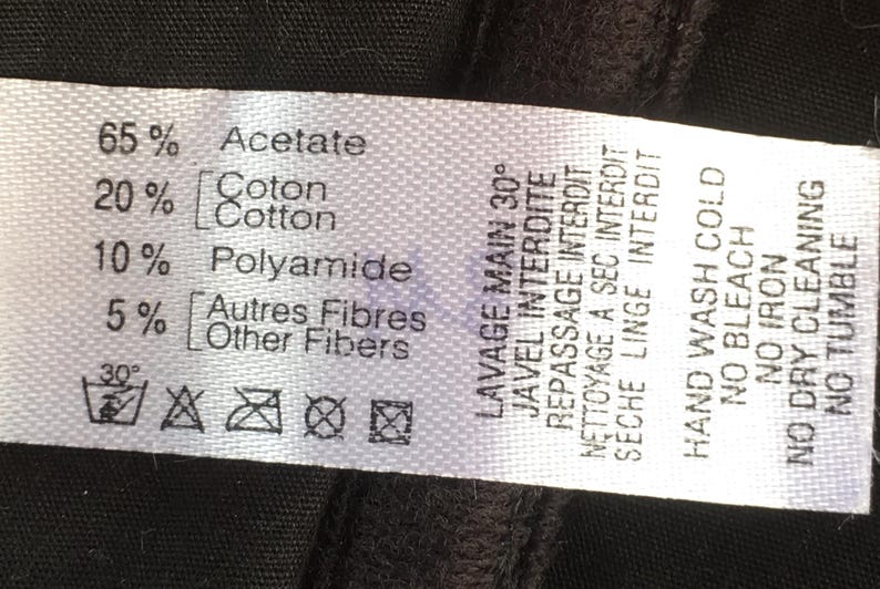 May include: A white fabric clothing label with black text detailing fabric composition: 65% Acetate, 20% Cotton, 10% Polyamide, and 5% Other Fibers. Washing instructions include hand wash cold, no bleach, no iron, no dry cleaning, and no tumble.