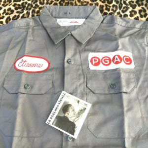 Op de afbeelding: Grijs werkhemd met twee rode patches. Eén patch zegt "Etienne" en de andere zegt "PGAC". Een klein wit kaartje met de tekst "LA DOUCHKA VINTAGE" is aan het shirt bevestigd.