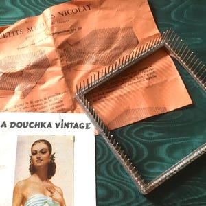Op de afbeelding: Een vierkant metalen weefraam met talloze rechtopstaande pinnen langs de omtrek, vergezeld van een gevouwen oranje instructieblad getiteld PETITS METIERS NICOLAY en een witte kaart met de tekst LA DOUCHKA VINTAGE en een portret van een persoon.