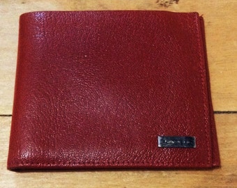 May include: A red leather wallet with a textured surface. The wallet has a rectangular shape and a small, silver-colored metal plate on the front. The wallet is closed and appears to be new.